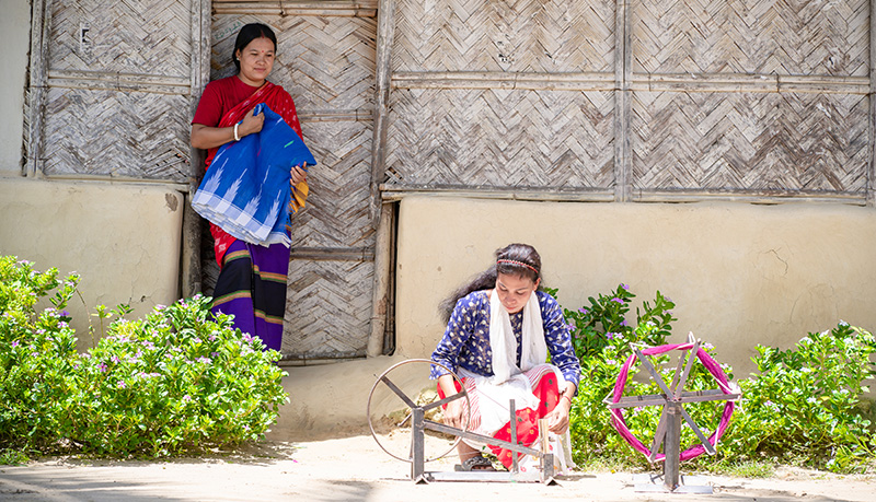 two women, one stooping with a wheeled device, in front of a dwelling